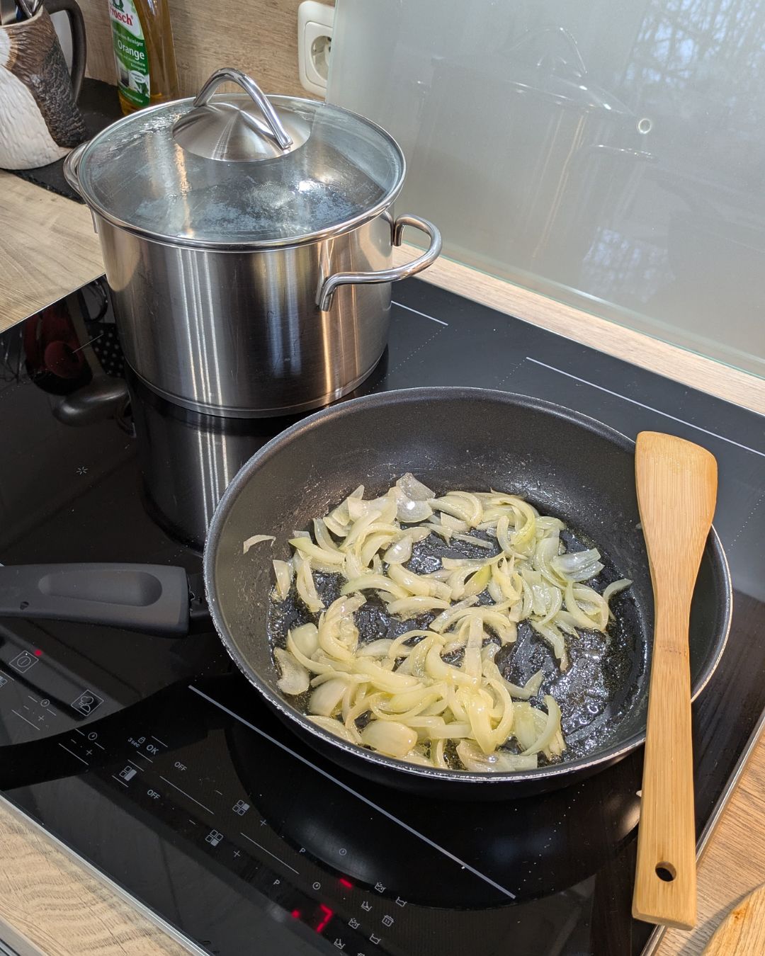 In Streifen geschnittene Zwiebeln brutzeln in einer Pfanne auf einem Induktionsherd, daneben steht ein großer Topf mit Nudelwasser.