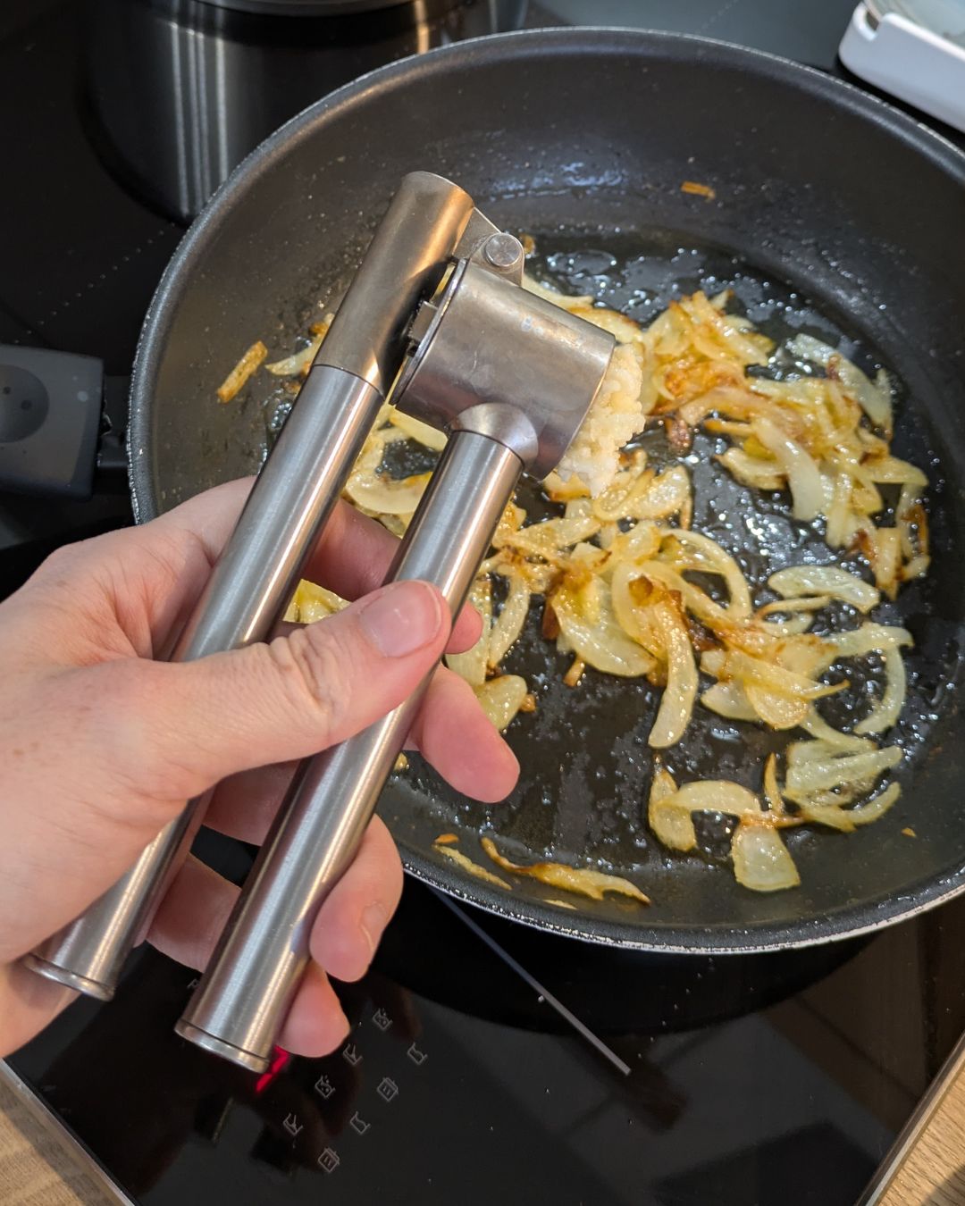 Eine Hand presst frischen Knoblauch mit einer Edelstahl-Knoblauchpresse direkt in die Pfanne zu den goldbraun gedünsteten Zwiebeln.