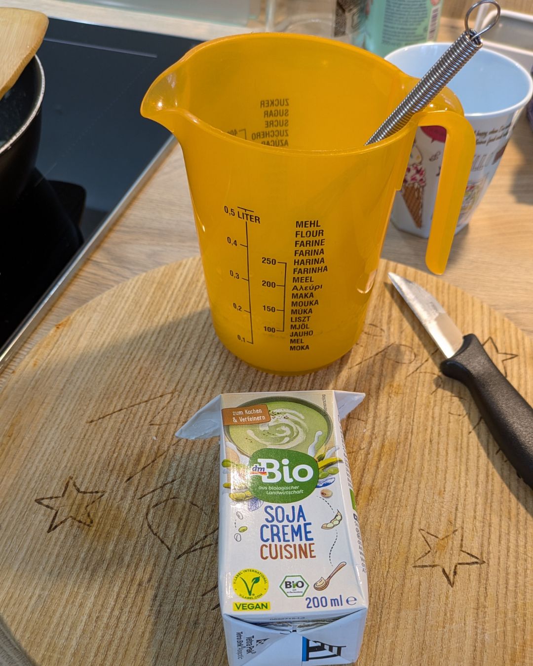 utaten für die Sauce: Ein gelber Messbecher und eine Packung vegane dm Bio Soja Creme Cuisine auf einem Holzschneidebrett.