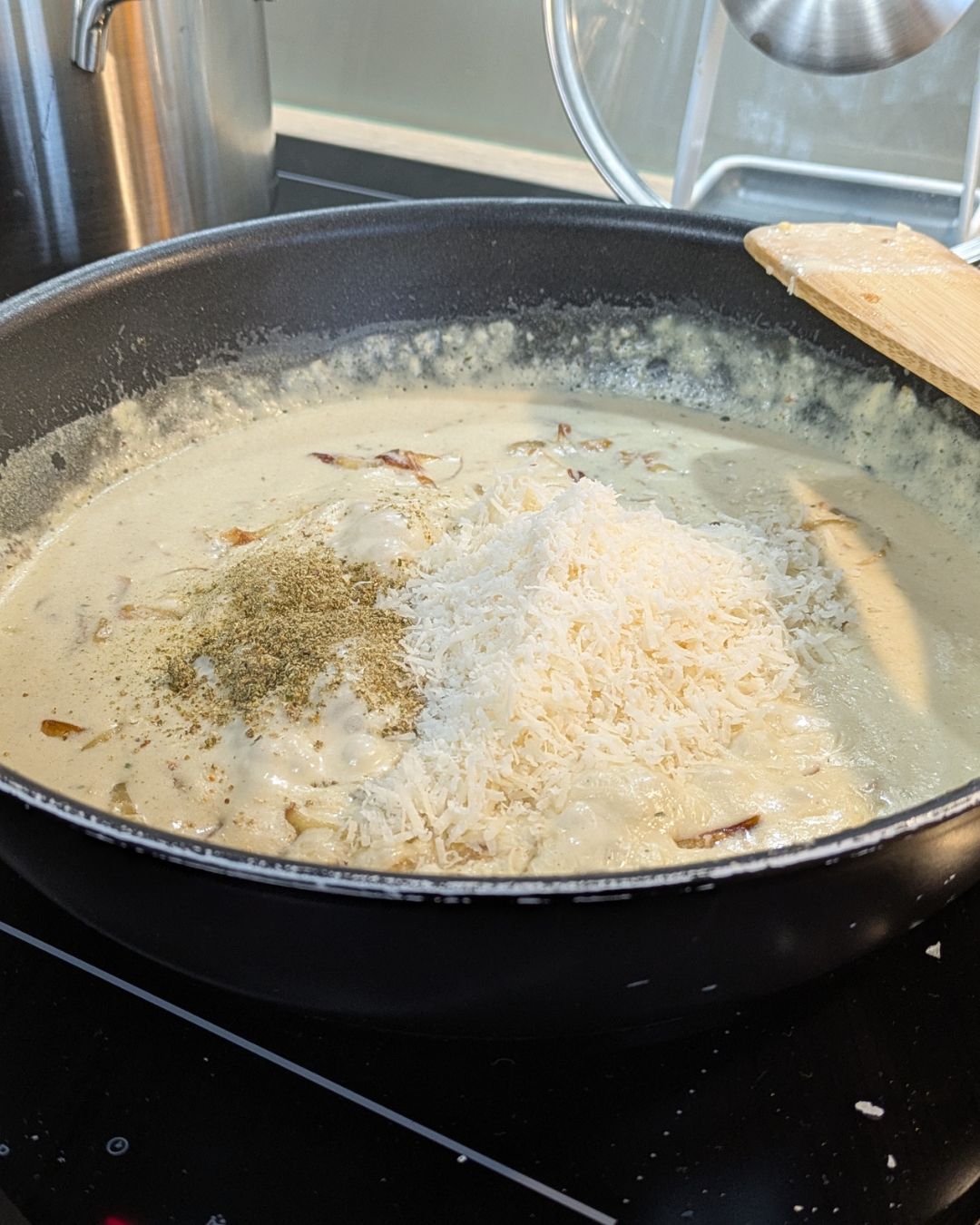 Die helle Pastasauce köchelt in der Pfanne, während geriebener Käse und Gewürze eingerührt werden.