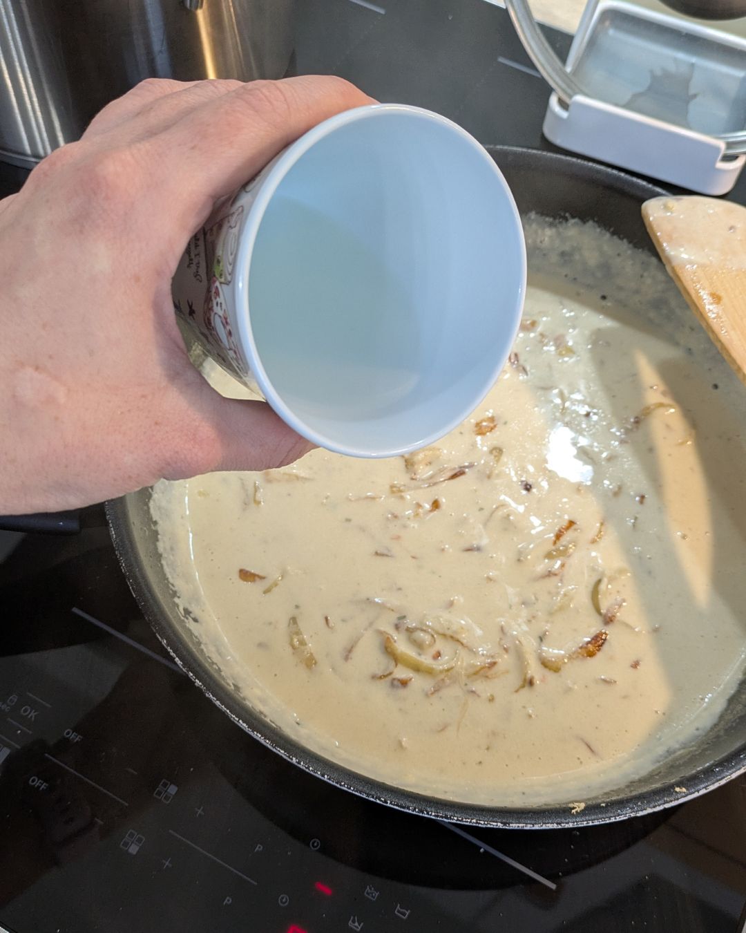 Eine Hand gießt einen Becher Wasser (oder Pastawasser) in die Pfanne zur cremigen Zwiebelsauce, um die Konsistenz zu verfeinern.