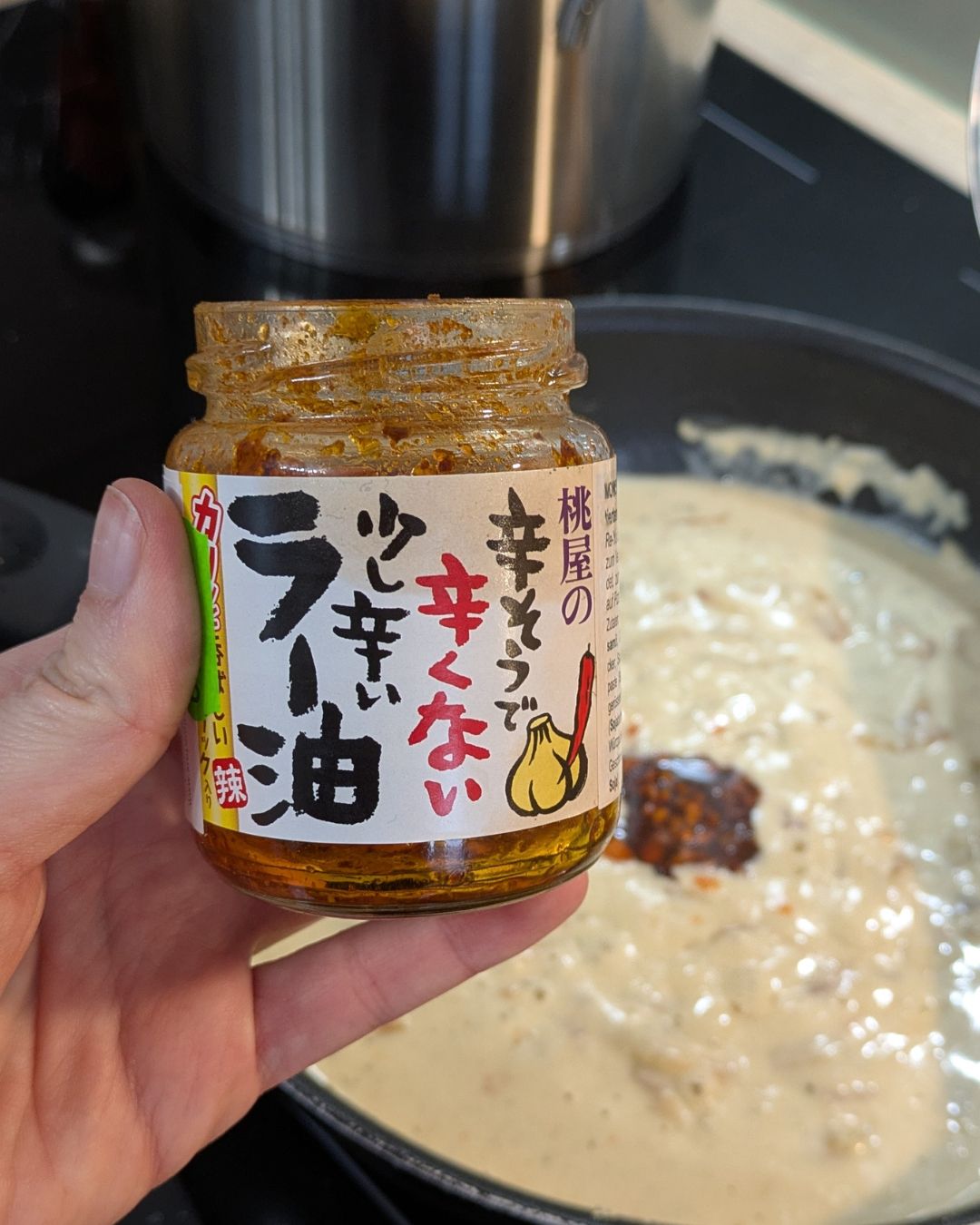 Nahaufnahme eines Glases japanisches Chili-Öl (Rayu) mit Knoblauch-Stückchen, das über die fertige helle Sauce gehalten wird.