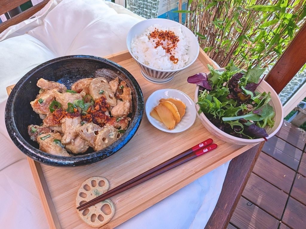 Vogelperspektive (Flat Lay) eines japanischen Tori Mayo Teishoku-Menüs auf einem Holztablett: Anordnung der Schalen mit Hähnchen-Lotuswurzel-Mix, Reis, Salat und eingelegtem Rettich. Die roten Essstäbchen ruhen auf einer Keramik-Lotuswurzel-Stäbchenablage, alles auf einem hellen Holztablett auf einer weißen Decke im Freien.