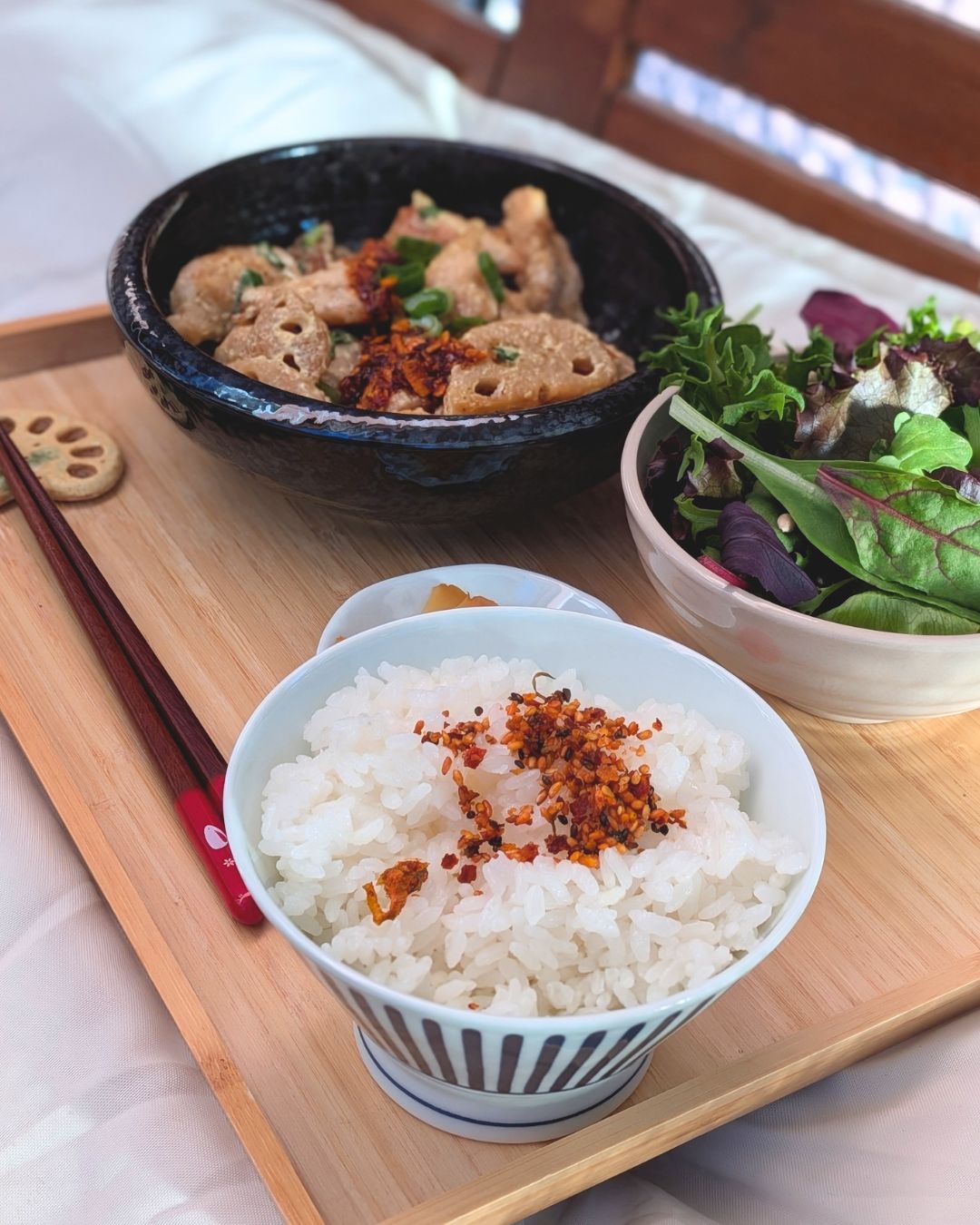 Traditionelles japanisches Teishoku-Menü mit Tori Mayo und Lotuswurzel in einer dunklen Schale, serviert auf einem Holztablett mit einer Schüssel japanischem Reis mit Furikake-Gewürz im Vordergrund, einem Beilagensalat und Essstäbchen.
