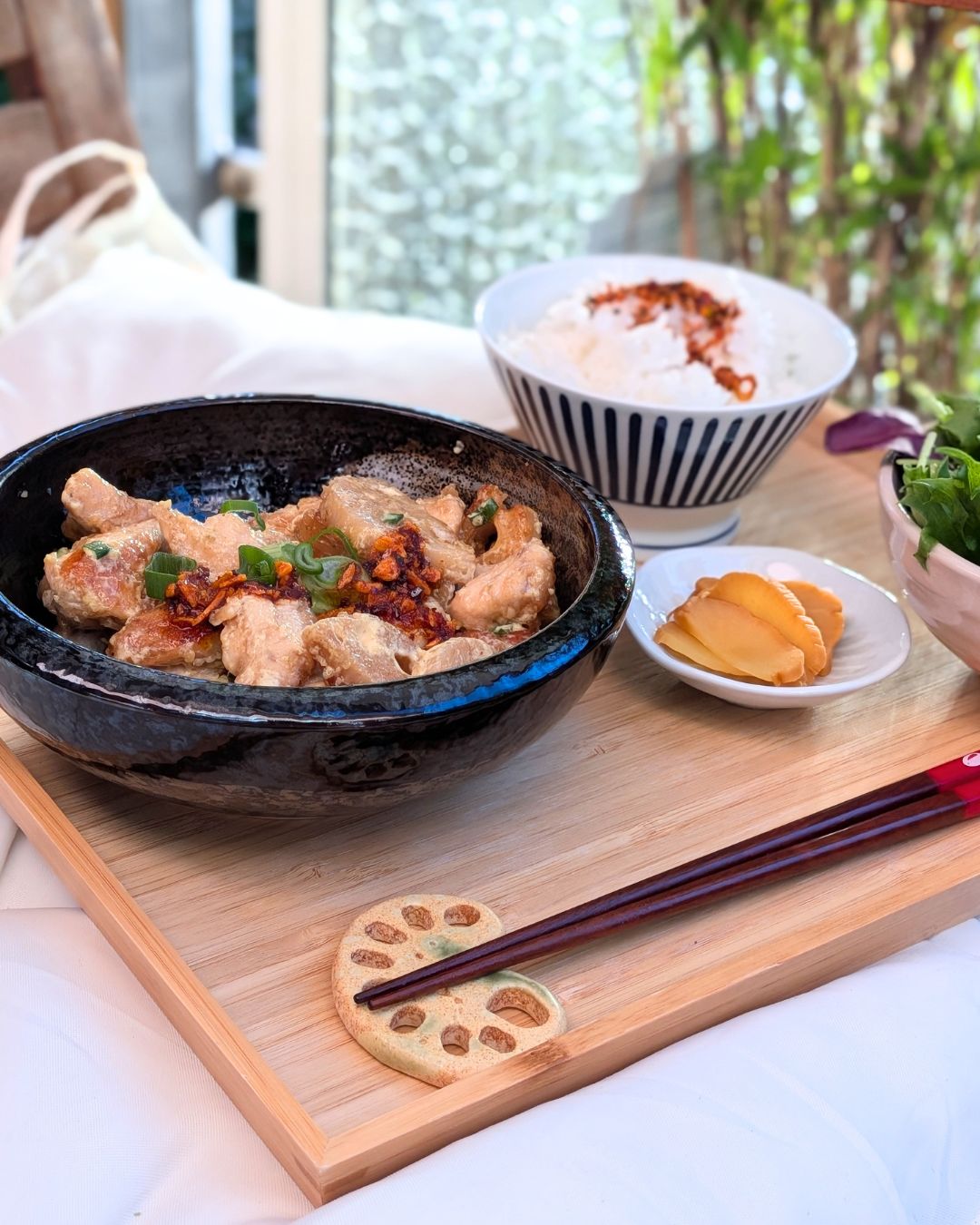 Japanisches Tori Mayo Rezept mit knusprigem Hähnchen und Lotuswurzel in einer schwarzen Keramikschale, garniert mit Frühlingszwiebeln und Chiliöl, serviert mit Reis, eingelegtem Gemüse (Tsukemono) und Essstäbchen auf einer Lotuswurzel-Stäbchenablage.