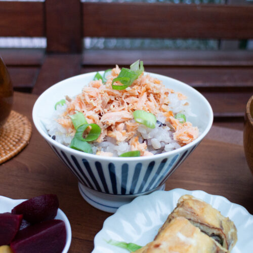 ine Mahlzeit im japanischen Stil: Eine blau-weiße Schüssel mit Reis, gekrönt mit Lachsflocken und Frühlingszwiebeln. Rote-Bete-Scheiben und Tamagoyaki stehen daneben.