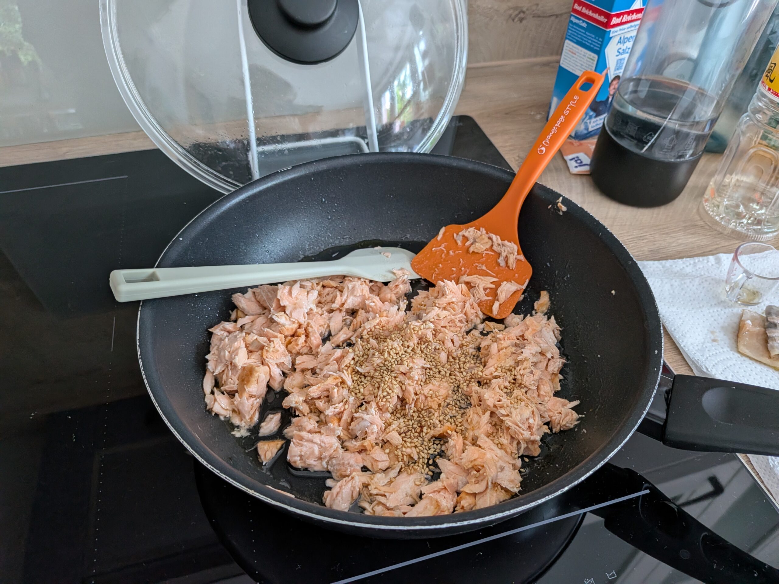 Eine Nahaufnahme der gekochten Lachsflocken in einer Bratpfanne auf einem Induktionskochfeld. Ein grüner und ein oranger Pfannenwender vermischen die Flocken. Sesam ist darüber gestreut.